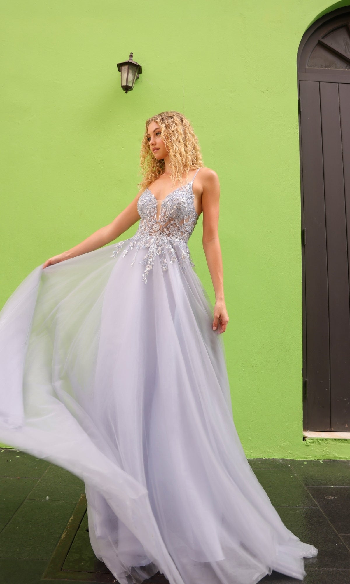 A woman in a Deep V-Back Long Tulle A-Line Prom Dress Q1391 stands before a vivid green wall with a black door and lamp, holding out the skirt of her light blue evening gown.